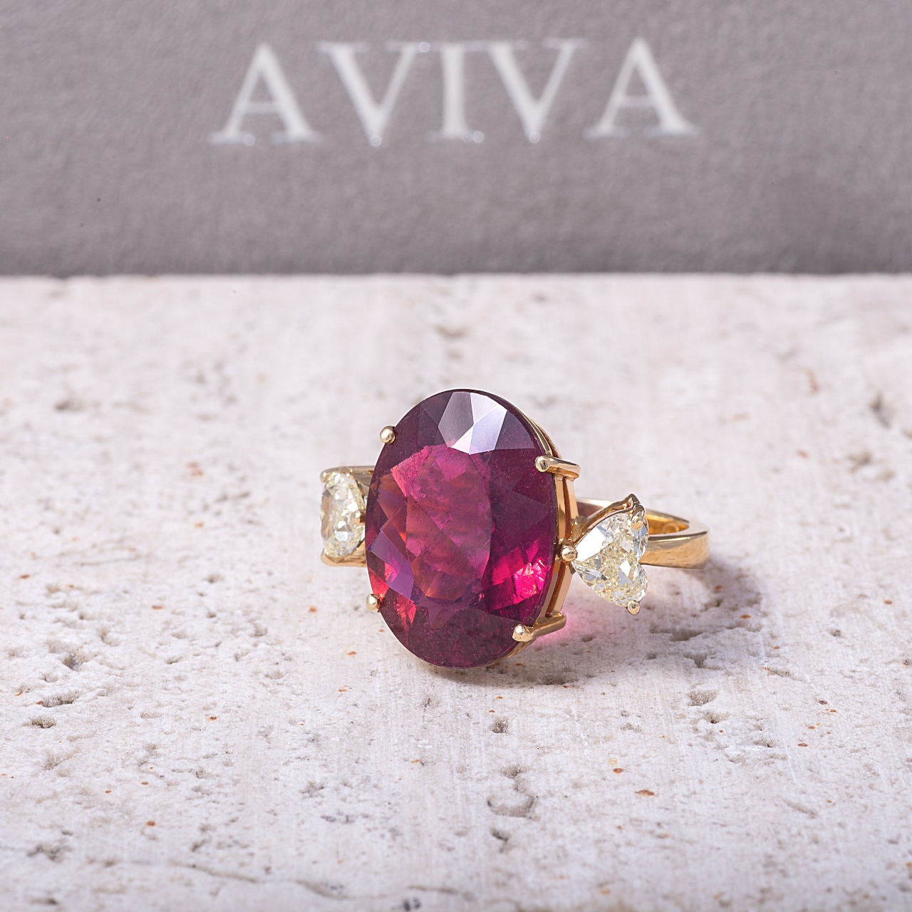 Anel de ouro com pedra rosa oval e dois diamantes, sobre a caixa da Aviva